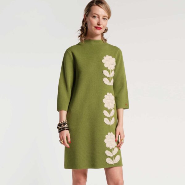 A woman in a grass green knit shift dress adorned with floral embroidery along the side.